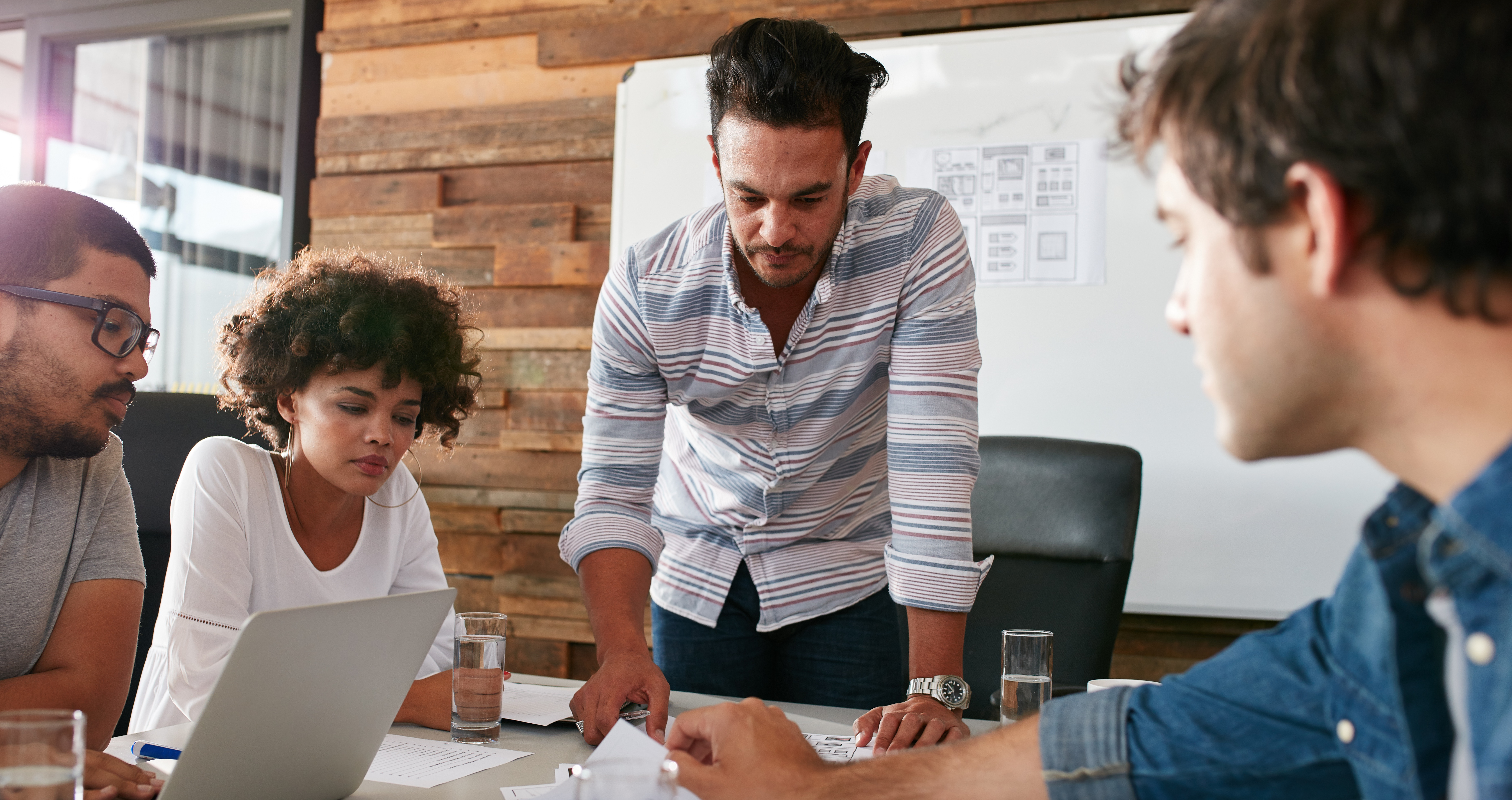 The image shows a photo of a marketing meeting taking place, with four team members around a table looking at information on paper and on a laptop.